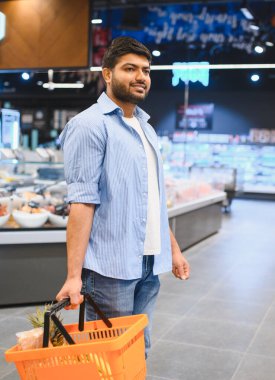 Marketin koridorlarında yürürken elinde alışveriş sepeti taşıyan genç Hintli bir adam.