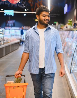 Market koridorunda yürürken elinde alışveriş sepeti taşıyan gülümseyen Hintli genç adam.