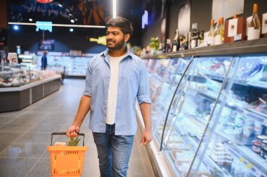 Genç Hintli bir adam bir alışveriş sepeti taşıyor ve süpermarketin soğutucu koridorlarında yürüyor, sebzeleri seçiyor.