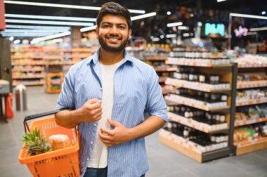 Genç Hintli adam bir süpermarkette alışveriş yaparken içinde ananas ve diğer malzemelerin olduğu bir alışveriş sepeti tutuyor ve gülümsüyor.
