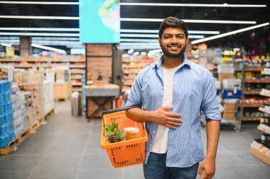 Genç Hintli adam elinde alışveriş sepeti tutarken gülümsüyor. Parlak ve düzenli bir süpermarket koridorunda bakkaliye dolu.