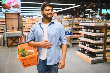 Market koridorunda yürürken alışveriş sepeti taşıyan genç Hintli adam, yiyecek satın alıyor.