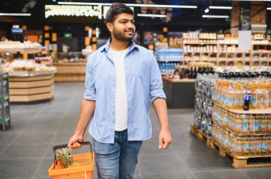 Market koridorlarında, içinde ananas bulunan alışveriş sepetiyle yürüyen genç Hintli adam, ürünleri seçiyor.