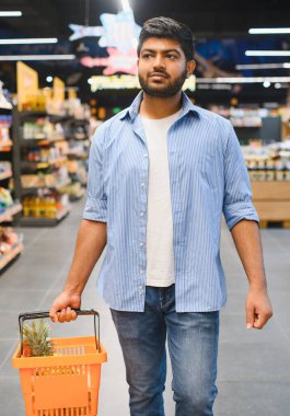 Bir süpermarketin koridorlarında yürürken elinde alışveriş sepeti taşıyan genç Hintli adam ürünleri seçiyor.