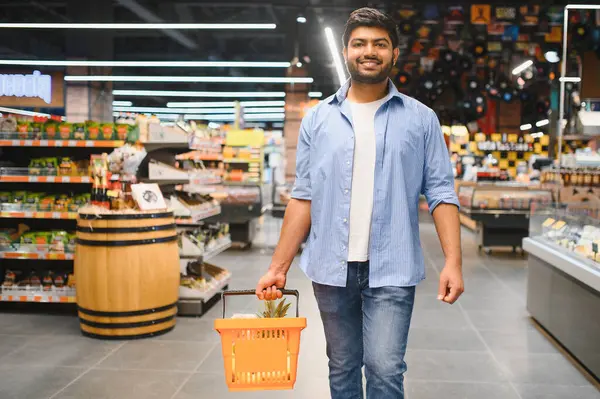 Genç Hintli bir adam bir alışveriş sepeti taşıyor. Parlak market koridorlarında yürüyor. Taze ürün ve ürünlere bakarken gülümsüyor.