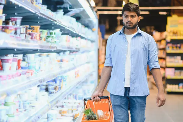 Genç Hintli bir adam markette dondurulmuş süt ürünleri reyonunda yürürken alışveriş sepsiyonu taşıyor.