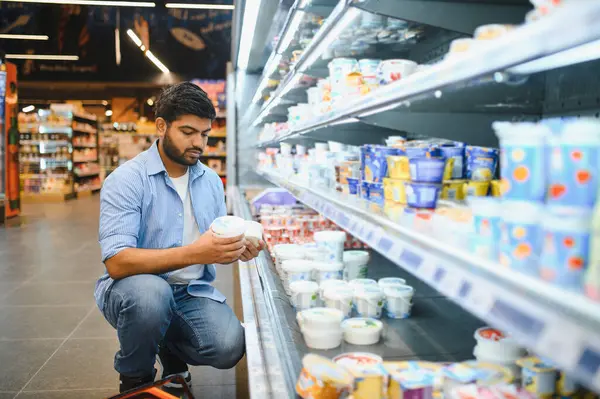 Yoğunlaşmış Hintli genç bir adam süpermarkette çömelmiş ve süt ürünlerini seçiyor.