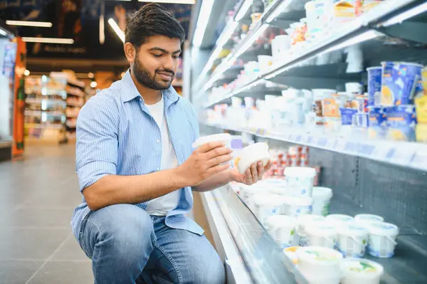 Konsantre olmuş genç Hintli adam bir markette soğutulmuş süt ürünleri reyonunun yanında çömelmiş iki yoğurt konteynırını karşılaştırıyor.