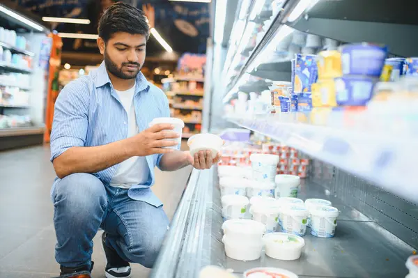 Genç Hintli bir adam yoğurt markalarını karşılaştırırken modern bir marketin dondurulmuş süt ürünleri reyonunda çömeliyor.