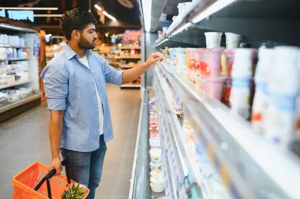 Ananaslı bir alışveriş sepeti tutan, parlak ışıklı bir süpermarkette soğutulmuş raflardan yoğurt seçen odaklanmış genç Hintli bir adam.