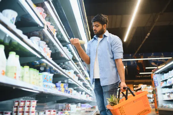 Genç Hintli bir adam süpermarkette alışveriş yapıyor, bir ürün etiketi okuyor, içinde ananas olan bir alışveriş sepeti tutuyor, süt ürünleri bölümünde.