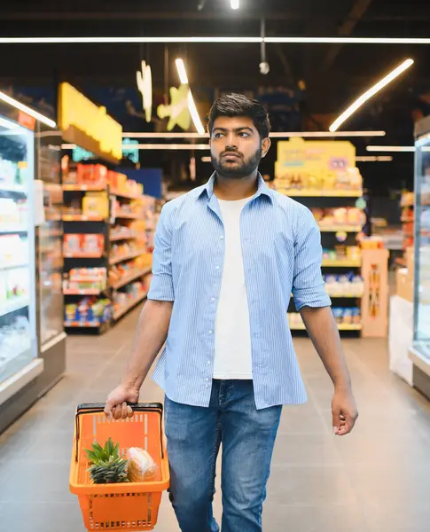 Genç Hintli bir adam elinde bir alışveriş sepeti, parlak, organize bir market koridorunda yürürken, düşünceli bir şekilde taze ürünleri seçiyor.