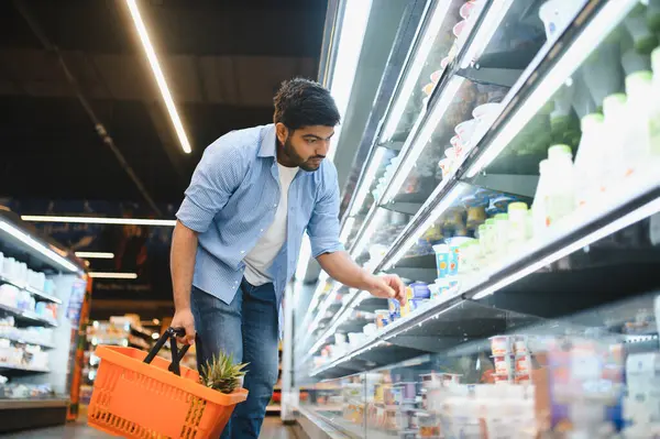 Odaklanmış müşteri dondurulmuş raflardan yoğurt seçiyor, ananaslı alışveriş sepeti tutuyor, modern süpermarkette market alışverişi yapıyor.