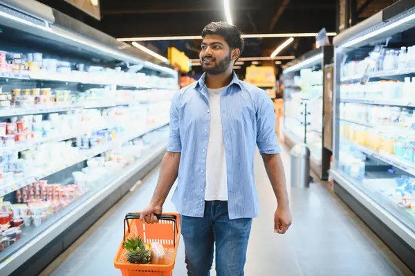 Müşteri, açık, modern bir süpermarkette alışveriş yaparken içi yiyecek dolu bir sepet taşıyor, taze ürünlere ve diğer ürünlere göz atıyor.