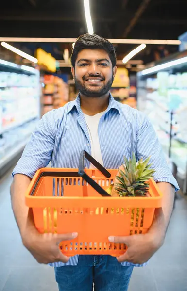 Hintli bir adam bakkalda içinde ananas ve muz bulunan portakal dolu bir alışveriş sepeti tutarken gülümsüyor.