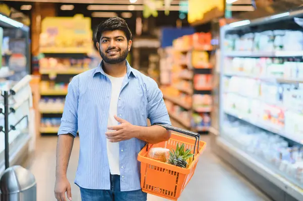 Genç Hintli bir adam elinde bir alışveriş sepeti, parlak ışıklı bir bakkal koridorunda yürürken, taze ürünler ve sebzeler seçiyor.