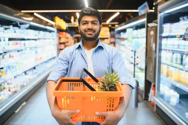 İçi ananas dolu turuncu bir alışveriş sepeti tutan gülümseyen Hintli müşteri, süpermarkette alışveriş yaparken.