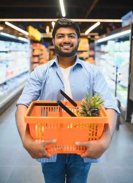Marketten alışveriş yaparken ananaslı turuncu bir alışveriş sepeti taşıyan mutlu Hintli müşteri.