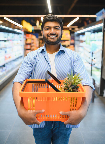 Счастливый индийский клиент с оранжевой корзиной с ананасом во время покупки продуктов в супермаркете