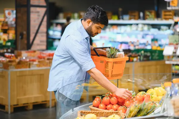 Genç Hintli adam taze ürün almak için alışveriş yapıyor, parlak ışıklı bir markette domates topluyor.