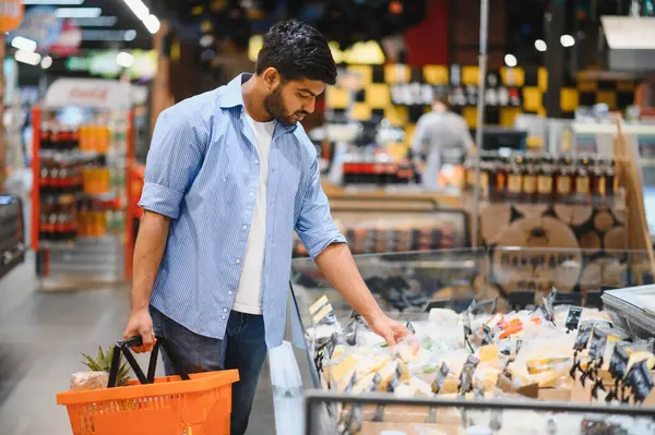 Müşteri marketin soğutulmuş bölümünden peynir seçiyor, elinde ekmek ve ananas ile alışveriş sepeti tutuyor.