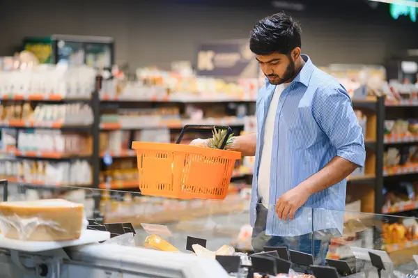 Müşteri marketteki soğutucudan peynir seçiyor, elinde turuncu alışveriş sepeti var.