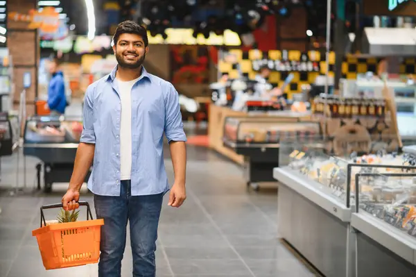 Genç Hintli bir adam alışveriş sepeti taşırken gülümsüyor ve hareketli bir marketin parlak koridorlarında yürüyor.