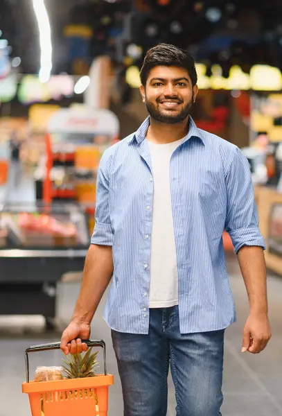 Genç Hintli bir adam alışveriş sepeti taşıyor. İçi taze yiyeceklerle dolu. Canlı süpermarket koridorlarında dolaşırken mutlu bir şekilde gülümsüyor.