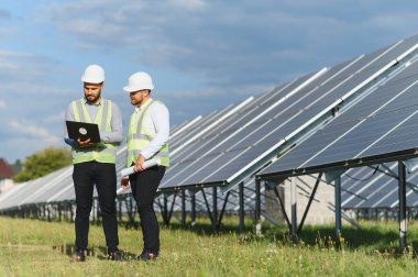 Güvenlik miğferleri ve yeleklerindeki iki mühendis yenilenebilir enerji ve modern teknolojiyi sembolize eden bir dizüstü bilgisayarla güneş panellerini inceliyor.