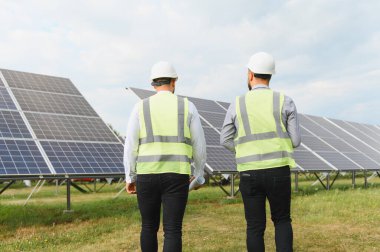 Güvenlik yelekleri ve miğferler üzerinde çalışan iki mühendis, yenilenebilir enerji ve temiz teknolojiyi sembolize eden yeşil alandaki büyük güneş panellerini inceliyor.