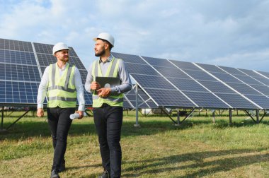 Güvenlik yelekleri ve miğferler giyen iki erkek mühendis, temiz enerji ve sürdürülebilirliği sembolize eden bir alan alanındaki güneş panellerini inceliyor.