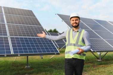 Kasketli ve güvenlik yeleği giymiş gülümseyen adam elinde planlar, güneş enerjisi panellerinin önünde duruyor.