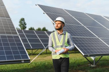 Güvenlik yeleği ve başlık takan mühendis güneş çiftliğini planlarla inceliyor ve temiz yenilenebilir enerji üretimine odaklanıyor.