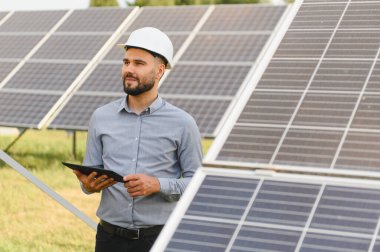 Sert bir şapka takan genç adam, güneşli bir alanda yenilenebilir enerji güneş panellerini kontrol ediyor, elinde dijital bir tablet tutuyor.