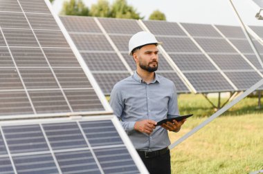 Genç mühendis sabit bir şapka takıyor ve elinde bir tablet tutuyor. Bir elektrik santralinde güneş panellerini inceliyor. Sürdürülebilir enerji kavramı