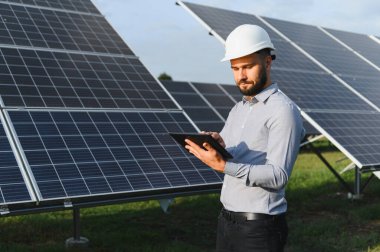 Güneş enerjisi çiftliğinde çalışan, sürdürülebilirlik için dijital tabletteki yenilenebilir enerji verilerini kontrol eden, sıkı şapkalı bir adam.