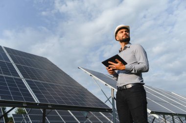 Erkek mühendis, başlık takıyor ve elinde tablet tutuyor. Güneş enerjisi santralindeki fotovoltaik panelleri kontrol ediyor.
