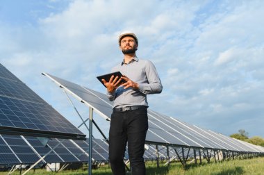 Baret takan ve elinde tablet tutan mühendis yenilenebilir enerji üretimi için güneş paneli alanını inceliyor.