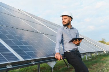 Başlık takan mühendis tablet tutuyor, güneş panellerinin yanında duruyor, sürdürülebilir teknoloji için enerji santrali performansını izliyor.