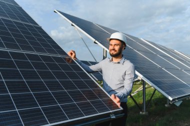 Sert şapkalı erkek mühendis, güneşli ve yenilenebilir enerji çiftliğindeki güneş panellerini incelerken gülümsüyor. Temiz teknolojik ilerlemeyi simgeliyor.