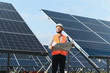 Güneş enerjisi santralinde çalışan erkek mühendis elinde bir proje tutuyor, sürdürülebilirlik için yenilenebilir enerji üretimini analiz ediyor.