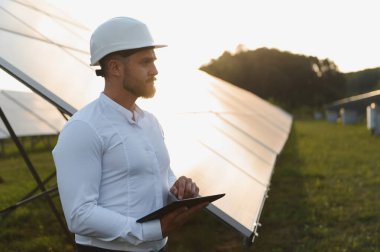 Günbatımında güneş panelleri sıralarını denetleyen sert şapkalı bir adam yenilenebilir enerji sistemini izlemek için dijital tablet kullanıyor.