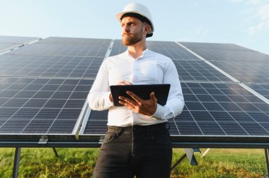Dijital tablet kullanan, güneşli bir günde güneş panellerinin önünde duran sert şapka ve beyaz gömlek giyen mühendis.