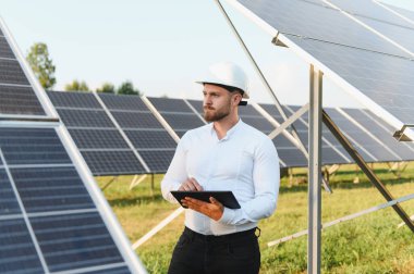 Dijital tablet tutan ve bir elektrik santralinde güneş panellerini denetleyen sert şapka ve beyaz gömlek giyen genç bir mühendis.