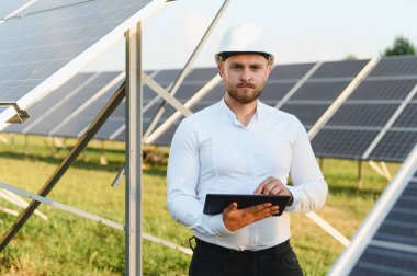 Elinde tablet olan sakallı adam, büyük bir güneş enerjisi santralinde sistem izleme ve bakımını yapıyor.