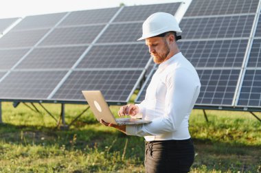 Sert şapka takan mühendis güneş panelinin performansını kontrol ediyor ve güneş enerjisi santralindeki bir laptopta veri analiz ediyor.