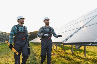 Sert şapka ve üniforma giyen iki mühendis güneş panellerini inceliyor, sürdürülebilir enerji çözümleri planlıyorlar.