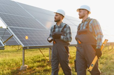 Güneş paneli alanını inceleyen, gün batımında kurulum ve sürdürülebilir enerji gelişimini tartışan sert şapkalı mühendisler