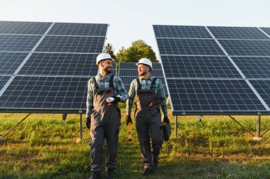 Bir güneş enerjisi santralinde yürüyen mühendisler, ellerinde planlar ve araçlar, sürdürülebilirlik ve yeşil enerji üzerinde işbirliği yapıyorlar.
