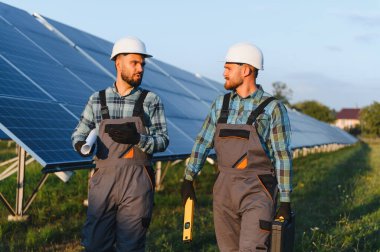 İki erkek mühendis, büyük bir güneş enerjisi santralinde çalışıyorlar, ellerinde belgeler ve aletler var.